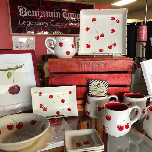 Display of cherry-themed pottery, including plates, mugs, and a pitcher, set against a rustic wooden backdrop.