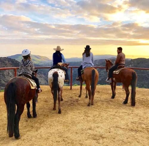 Four people on horseback overlooking a scenic landscape at sunset, with mountains and clouds in the background.