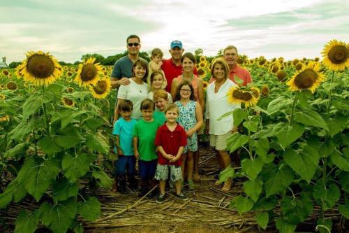 A group of people posing together in a sunflower field, surrounded by tall sunflowers under a cloudy sky.