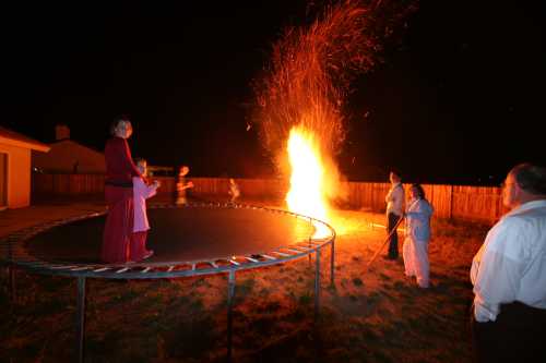 A group of people near a trampoline watch a large fire at night, with sparks flying into the air.