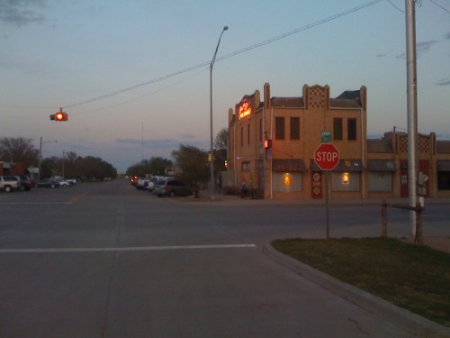 A street corner at dusk featuring a stop sign, traffic light, and a brick building with a neon sign.