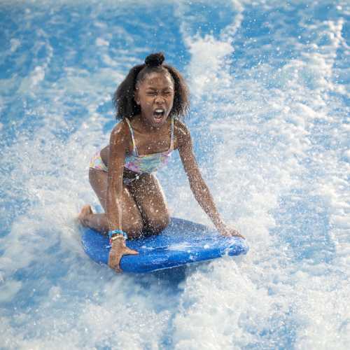 A young girl rides a blue boogie board on a wave, splashing water around her as she enjoys the ride.