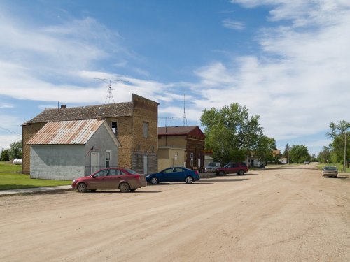 A quiet dirt road lined with buildings and parked cars under a blue sky with scattered clouds.
