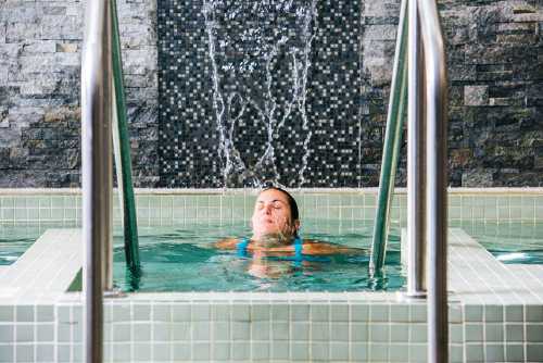 A woman relaxes in a spa pool, water cascading down a stone wall behind her.