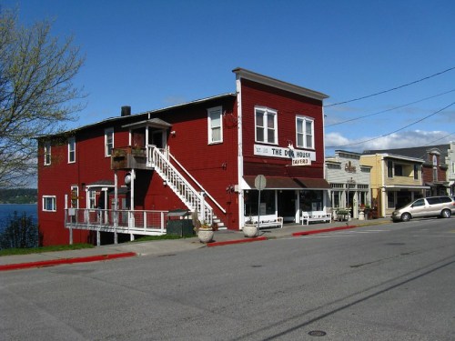 A red two-story building labeled "The Old House" near a waterfront, with nearby shops and clear blue skies.
