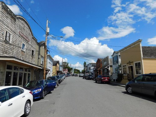 A quaint street lined with parked cars and charming buildings under a blue sky with fluffy clouds.