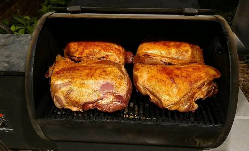 Four large pieces of meat seasoned and smoking on a grill, with a smoky atmosphere and greenery in the background.