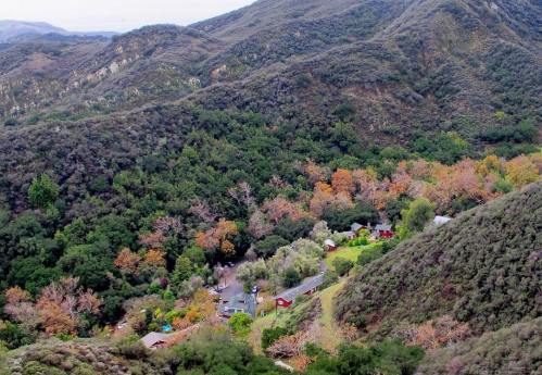 Aerial view of a lush valley with a small village, surrounded by rolling green hills and autumn-colored trees.
