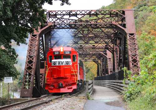 A red train crosses a vintage iron bridge surrounded by autumn foliage. Smoke billows from the locomotive.