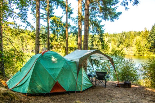 A green tent and canopy set up by a serene lake, surrounded by tall trees and lush greenery.