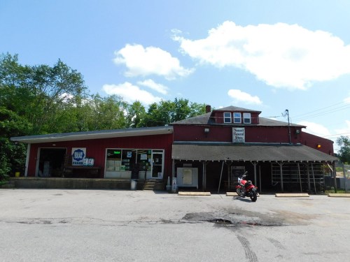 A red building with a porch and a motorcycle parked outside, surrounded by trees and a blue sky with clouds.