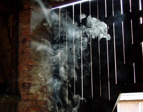 Smoke billows through gaps in a wooden wall, creating a mysterious atmosphere in a dimly lit space.