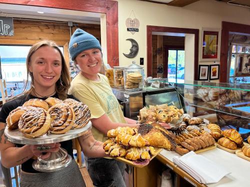 Two smiling bakers hold trays of assorted pastries in a cozy bakery, showcasing a variety of baked goods.