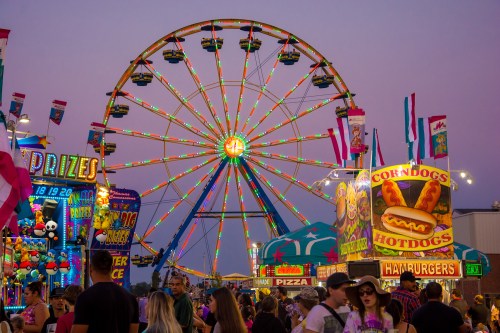 A vibrant carnival scene with a lit Ferris wheel and colorful food stalls, bustling with people at dusk.