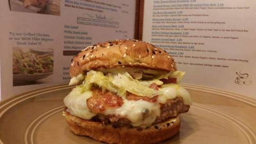 A sesame seed bun sandwich with a beef patty, melted cheese, lettuce, and sauce, served on a plate with a menu in the background.