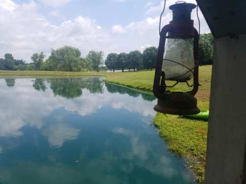 A rustic lantern hangs near a calm pond, reflecting trees and clouds in the water under a blue sky.
