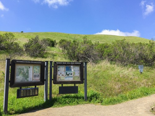 Two informational signs at a trailhead, with green hills and a blue sky in the background.