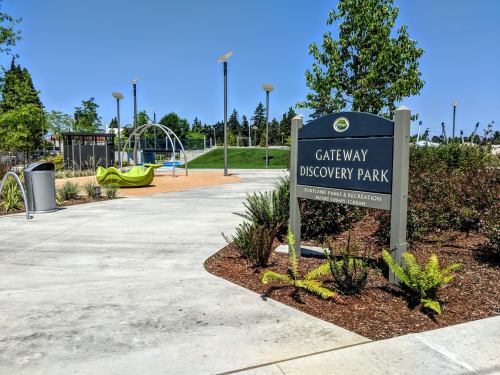 Sign for Gateway Discovery Park with playground equipment and greenery in a sunny outdoor setting.