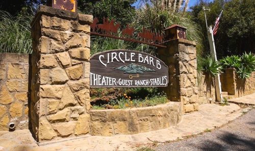 Sign for Circle Bar B Theater-Guest Ranch Stables, surrounded by greenery and a flagpole in the background.