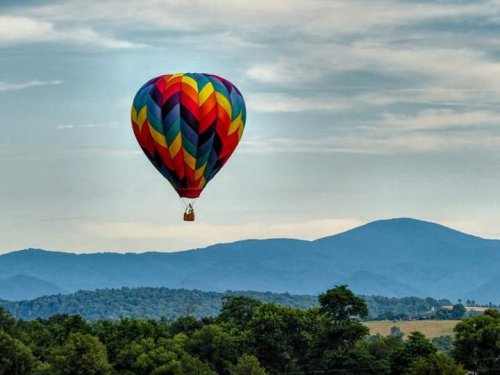 A colorful hot air balloon floats above green trees and rolling mountains under a cloudy sky.