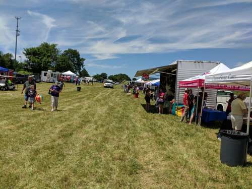 A sunny outdoor event with food stalls and vendors set up along a grassy field, with people walking around.