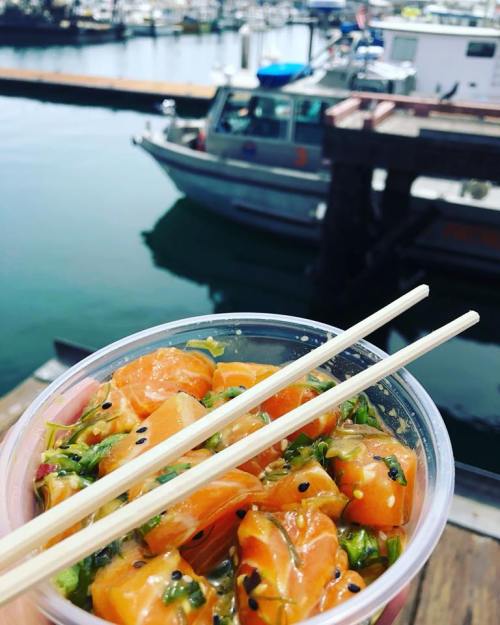 A bowl of salmon poke with chopsticks, held over a marina with boats in the background.