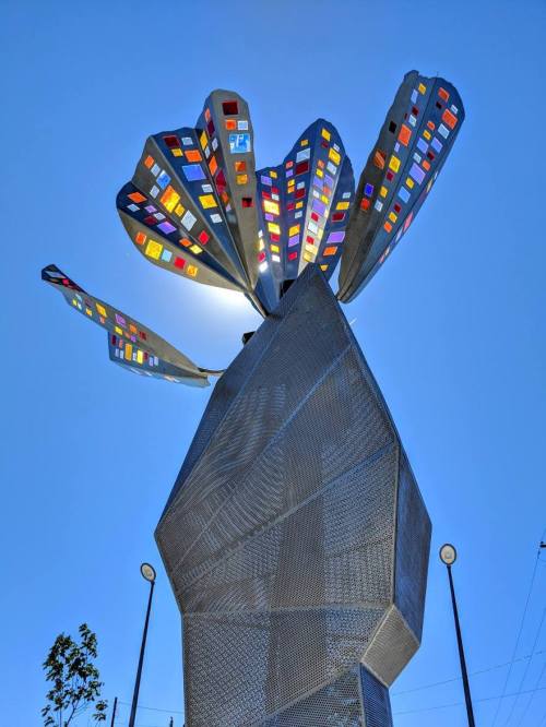 A large, metallic flower sculpture with colorful panels against a clear blue sky.