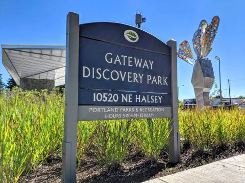 Sign for Gateway Discovery Park, featuring colorful butterfly sculpture and lush greenery, under a clear blue sky.