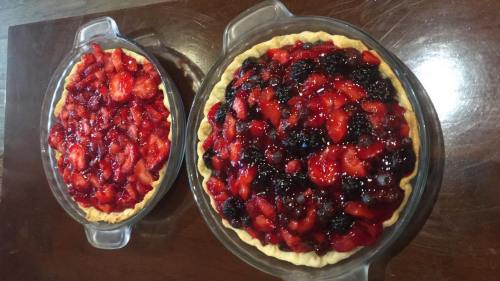 Two fruit pies on a wooden table, one filled with strawberries and the other with mixed berries, both in clear glass dishes.