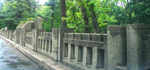 A weathered stone wall along a tree-lined path, surrounded by lush greenery and a wet pavement.
