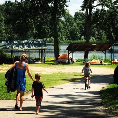 A woman and a child walk towards a lake, while another child rides a bike nearby. Tents are set up in the background.