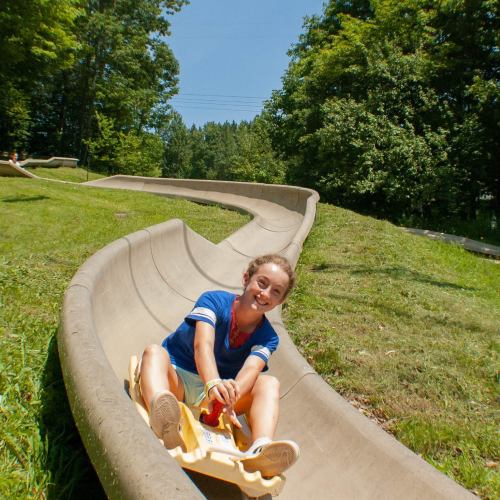 A child smiles while riding a sled down a winding concrete slide surrounded by green trees and grass.