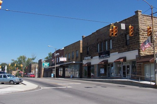 A sunny street view of a small town with shops, traffic lights, and parked cars along the road.