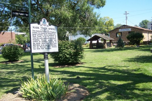 Sign for Mark Twain Spring in a park, with a gazebo and greenery in the background.