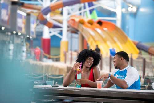 A couple enjoys colorful drinks at a water park, with water slides in the background.