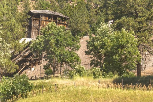 A weathered wooden structure on a hillside, surrounded by trees and grassy fields under a clear sky.