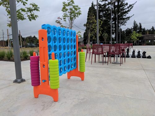A colorful outdoor connect-four game stands beside chairs and a large chess set in a park setting.