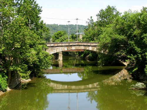A stone bridge arches over a calm river, surrounded by lush green trees and reflecting in the water below.