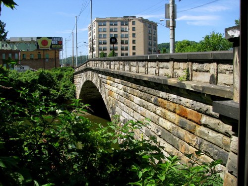 A stone arch bridge over a river, surrounded by greenery, with a building and blue sky in the background.