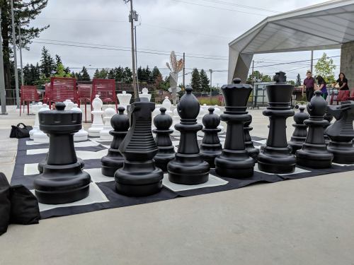 A large outdoor chess set with black and white pieces arranged on a checkered board, surrounded by spectators.
