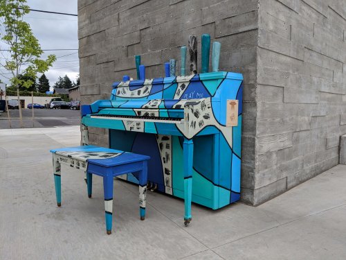 Colorful, artistic piano and bench with blue and white designs, placed against a textured gray wall outdoors.