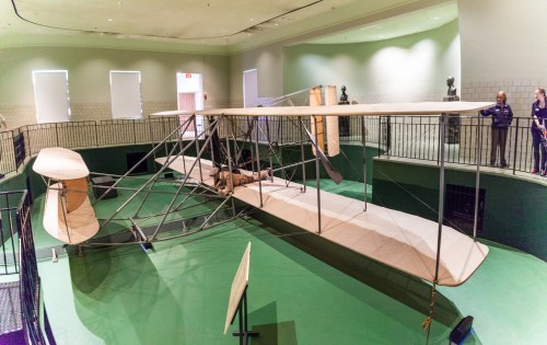 A vintage airplane model displayed in a museum, surrounded by visitors and green walls.