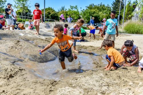 Children playing in a sandy area with water, enjoying a sunny day at a park.