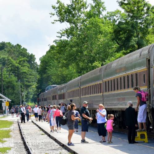 A busy train station with people boarding a vintage train surrounded by trees on a sunny day.