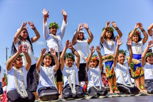 A group of children in traditional attire performing a dance, raising their hands, with floral crowns and smiles.