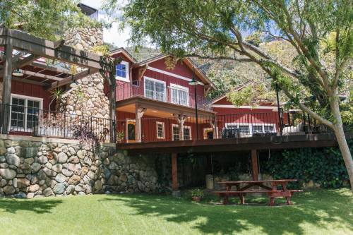 A red house with a stone foundation, surrounded by greenery and featuring a wooden deck and picnic table.