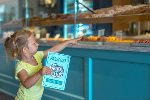 A young girl in a yellow shirt points at a display of donuts while holding a "Donut Trail" passport.