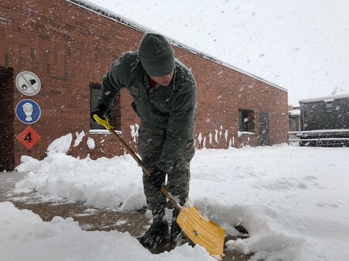 A person in military uniform shovels snow outside a building during a heavy snowfall.