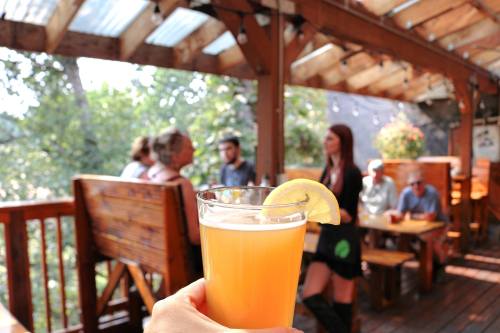 A hand holds a glass of beer with a lemon slice, set on a wooden deck with people socializing in the background.