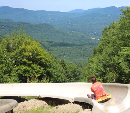 A child rides a summer slide down a hill, with lush green mountains in the background under a clear blue sky.